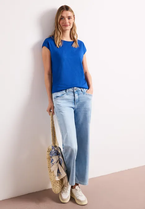 Vrouw leunt tegen witte muur, draagt blauw t-shirt, wijde spijkerbroek, witte sneakers en heeft een strozak.