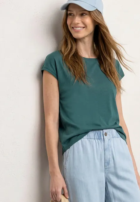 Een vrouw draagt een groen t-shirt, lichte spijkerbroek en een pet tegen een witte achtergrond.