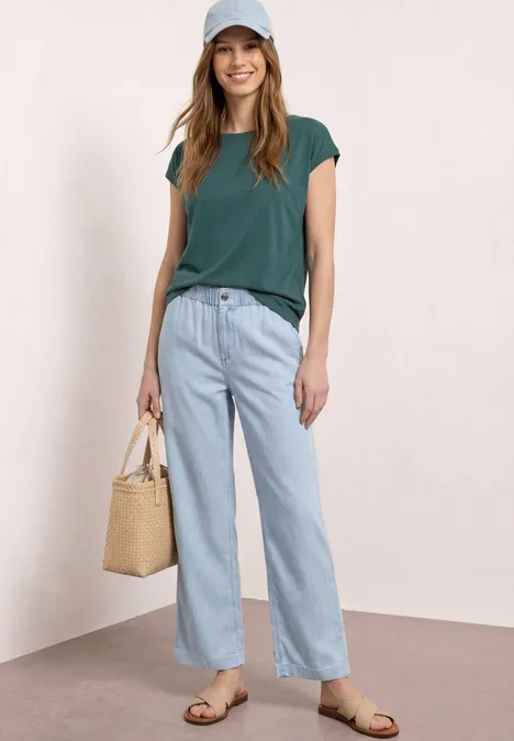 Vrouw in groen T-shirt, licht wijde jeans, sandalen en een strooien tas voor een neutrale achtergrond.