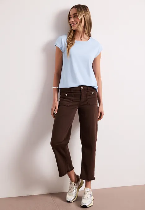 Vrouw in licht t-shirt, bruine 3/4-broek en sneakers voor een neutrale achtergrond.