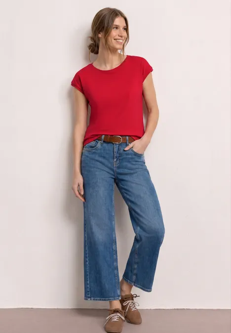 Vrouw voor lichte achtergrond, rood T-shirt, wijde jeans, bruine riem en schoenen.