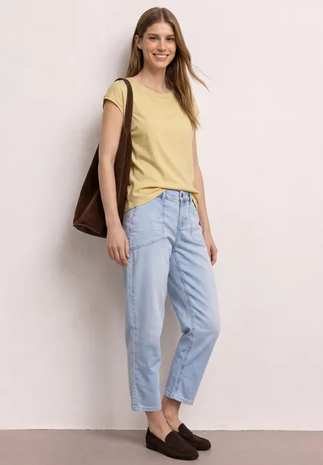 Frau in gelbem T-Shirt, hellblauen Jeans, braunen Loafern und brauner Tasche vor hellem Hintergrund.