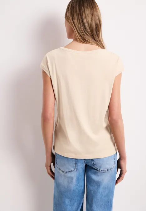 Vrouw gezien van achteren, draagt beige T-shirt met opgerolde mouwen en losse blauwe spijkerbroek.