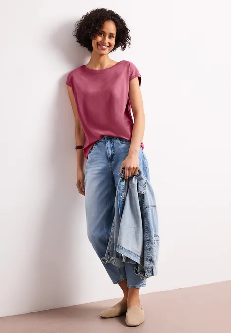 Vrouw staand in roze top en wijde jeans, houdt een lichtblauwe jeansjas in haar hand.