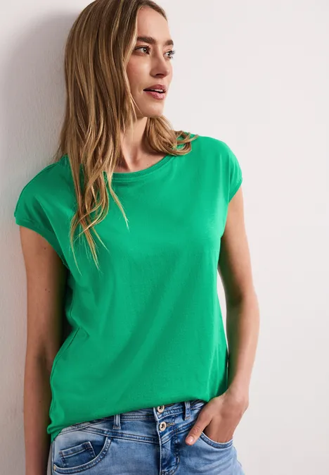 Vrouw in een groen t-shirt en blauwe spijkerbroek, met één hand in de zak, staand voor een witte muur.