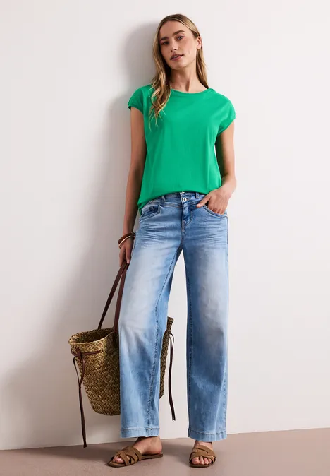 Vrouw met een groen t-shirt en wijde blauwe jeans, houdt een gevlochten strozak vast.