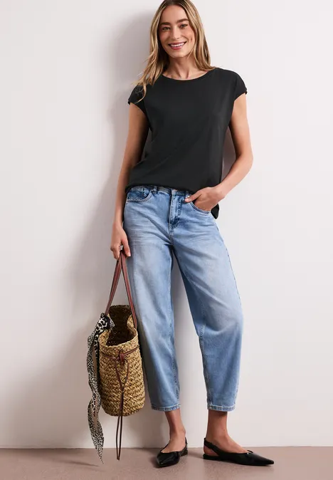 Een vrouw in een zwart T-shirt, blauwe jeans en zwarte ballerina’s met een rieten tas geleund tegen een muur.