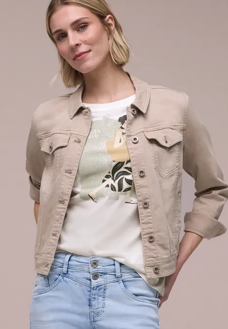 Vrouw in beige spijkerjack, wit T-shirt met bladprint en een lichtblauwe spijkerbroek.
