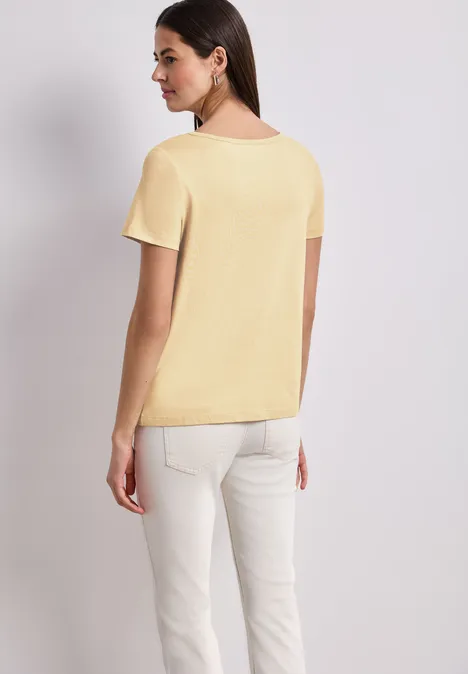 Vrouw van achteren in lichtgeel T-shirt en witte broek tegen lichte achtergrond.