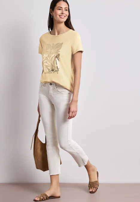 Vrouw in gele vlinderprint-T-shirt, witte driekwartbroek, bruine tas en luipaardprint sandalen.