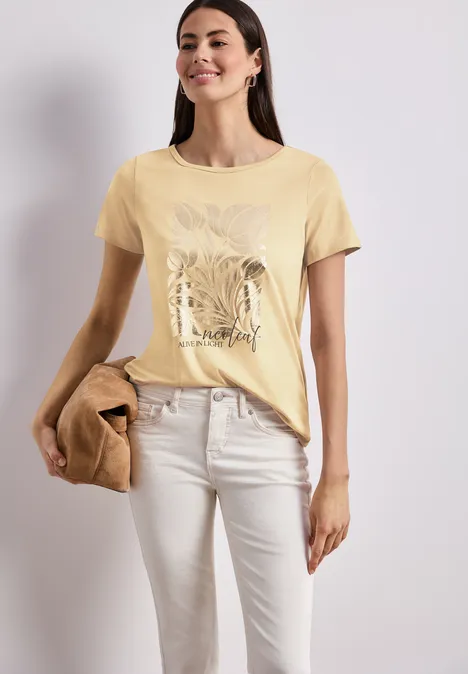 Vrouw met geel T-shirt met plantenprint, lichte broek en bruine tas tegen een effen achtergrond.