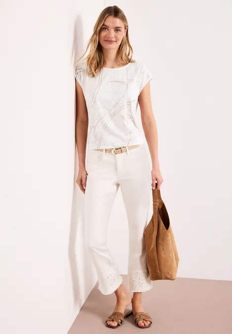Femme en haut en maille blanc, pantalon large blanc, sandales, sac marron, appuyée contre un mur blanc.