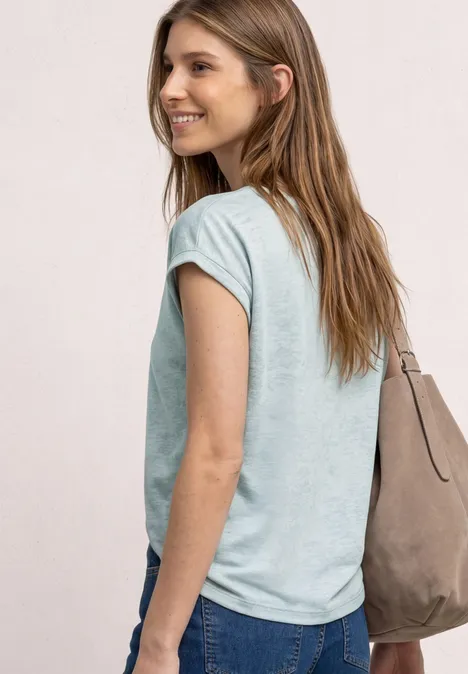 Femme de dos en t-shirt bleu clair, jean et sac beige à l’épaule sur fond neutre.
