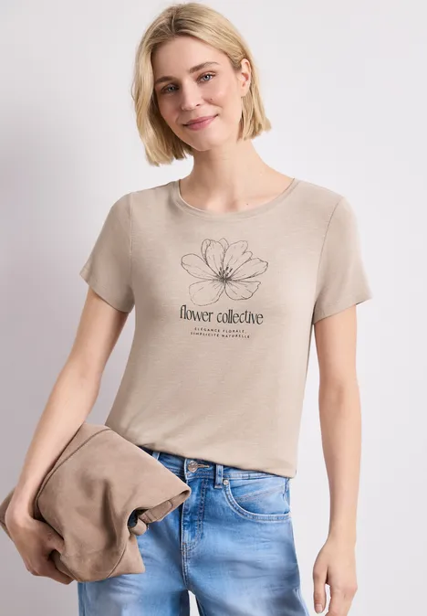 Frau in beigem T-Shirt mit Blumenmotiv und Jeans, hält passende Jacke in der Hand.