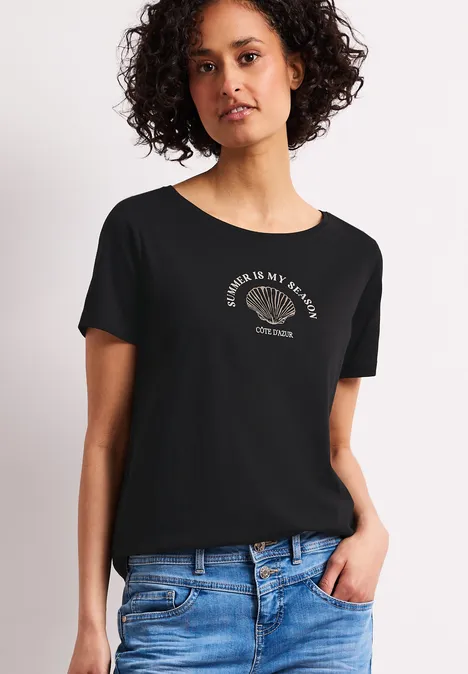 Vrouw draagt zwart T-shirt met tekst en blauwe jeans, staand voor een lichte achtergrond.