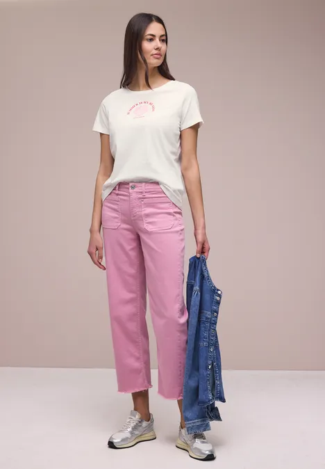 Shirt met schelpenprint off white Vrouw in wit T-shirt, roze jeans en sneakers, houdt een blauw spijkerjasje vast.