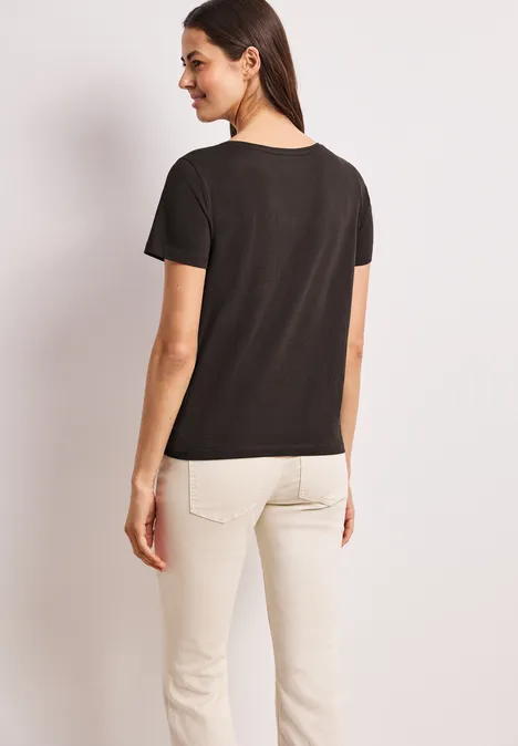 Frau von hinten in schwarzem T-Shirt und hellen Hosen vor neutralem Hintergrund.