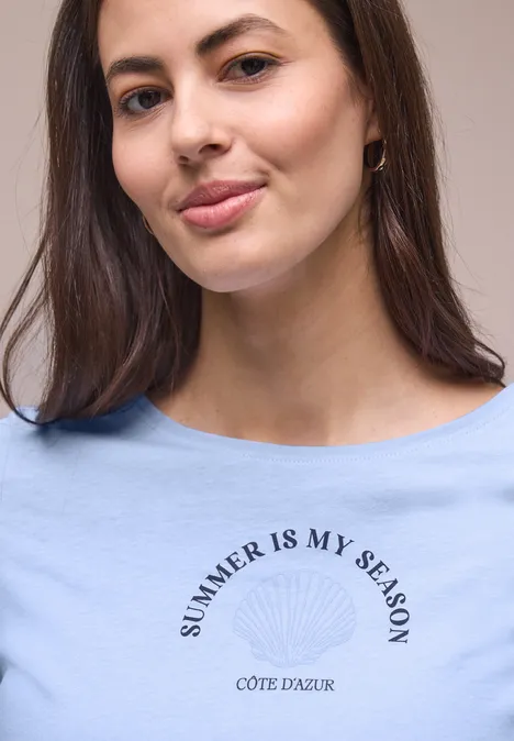 Shirt met schelpenprint cashmere blue Vrouw in blauw t-shirt met tekst SUMMER IS MY SEASON CÔTE D'AZUR, gezicht onscherp.