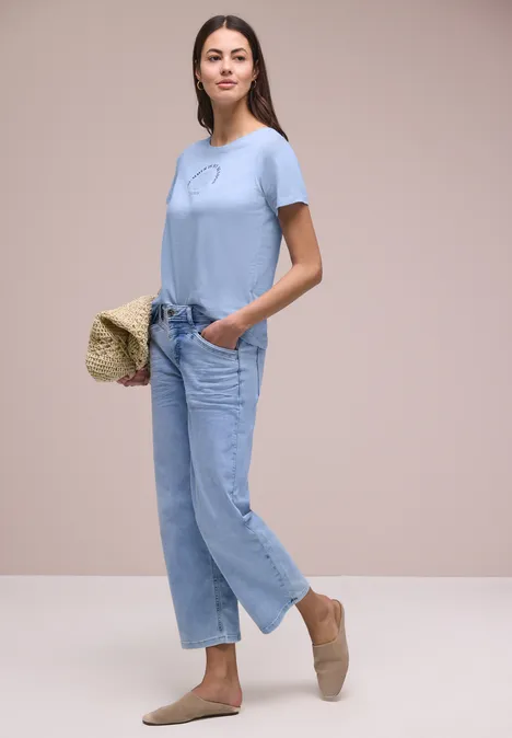 Shirt met schelpenprint cashmere blue Vrouw in lichtblauw T-shirt, losse jeans, beige espadrilles en een strooien tas.