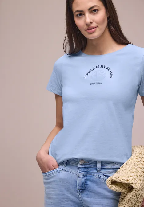 Shirt met schelpenprint cashmere blue Vrouw in lichtblauw shirt en jeans met strooien tas poseert.