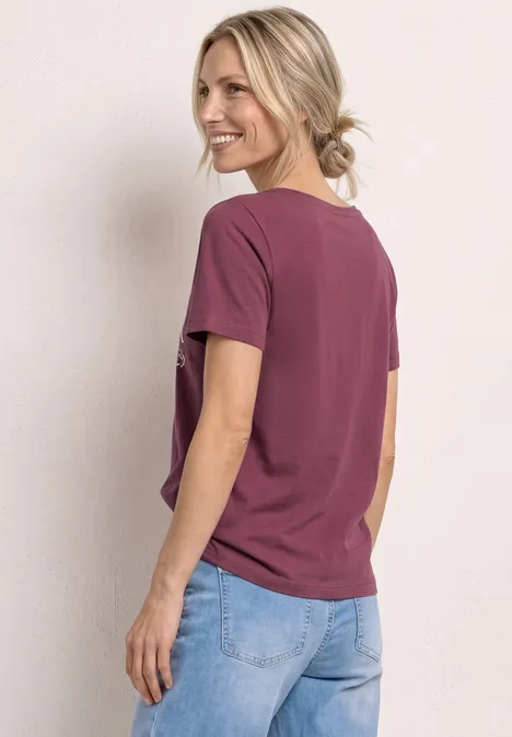 Persoon van achteren in kastanjebruin T-shirt en lichte spijkerbroek, zijwaarts kijkend, haar los in staart.