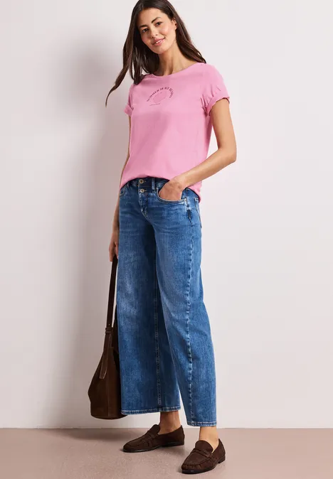 Vrouw in roze T-shirt, wijde cropped jeans en bruine loafers met een bruine schoudertas.
