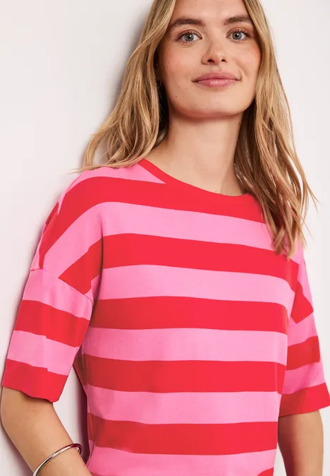 Shirt met ronde hals en strepen salsa red Jonge vrouw lacht, draagt roze-rood gestreept T-shirt met korte mouwen tegen lichte achtergrond.