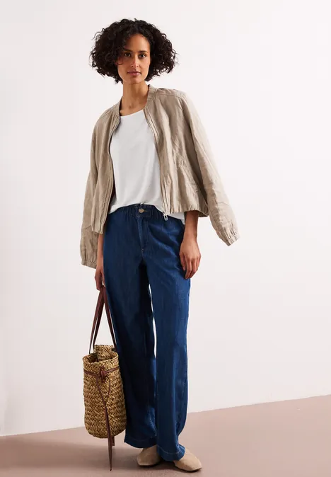 Vrouw in wit T-shirt, beige jasje, wijde jeans, gevlochten rieten tas en beige loafers.