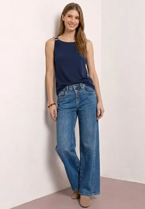 Vrouw leunt tegen muur, donkerblauw hemdje, wijde jeans en beige enkellaarsjes voor een strakke achtergrond.