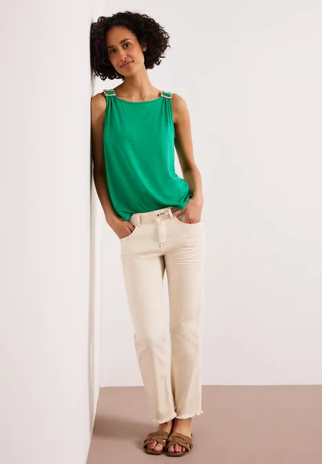 Iemand leunt tegen een witte muur, draagt een groen topje, beige broek en sandalen.