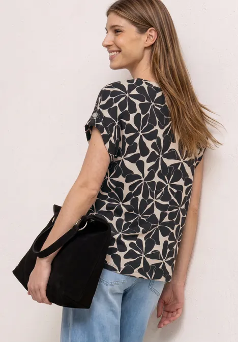 Frau mit langem blonden Haar trägt ein kurzärmeliges schwarz-weißes Blumenmuster-Shirt, Jeans und schwarze Tasche, von hinten.