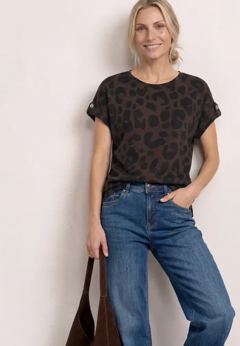 Vrouw in casual outfit: luipaardshirt, blauwe jeans en bruine tas tegen witte achtergrond.