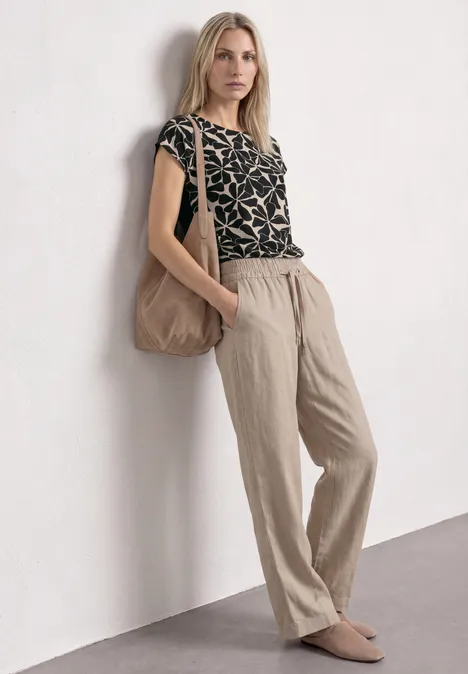 Vrouw leunt tegen muur, draagt beige linnenbroek, top met patroon, grote tas