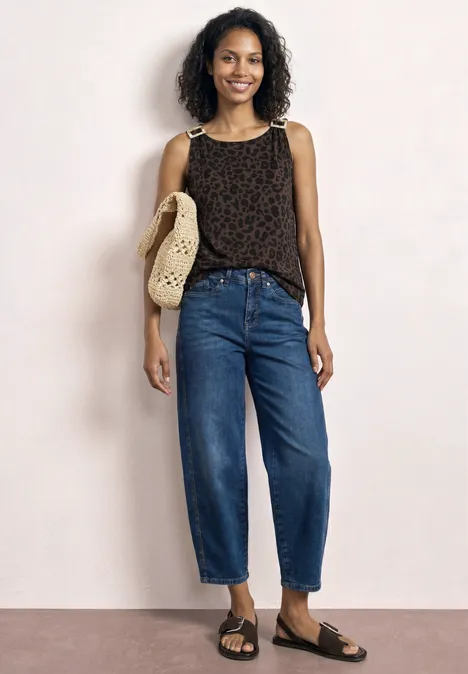 Frau trägt gemustertes Top, weite Jeans, flache Sandalen und eine Strohtasche vor hellem Hintergrund.