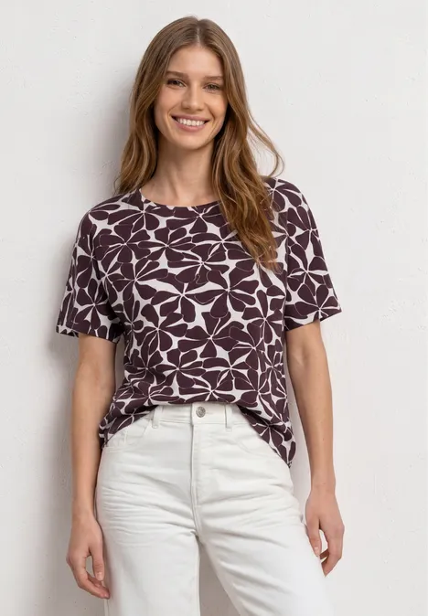 Frau in lila-weiß gemustertem T-Shirt und weißen Hosen vor hellem Hintergrund.