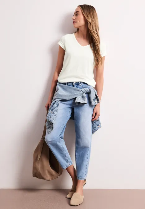 Vrouw in casual outfit met wit T-shirt, blauwe spijkerbroek, jas om de taille, beige tas en instappers