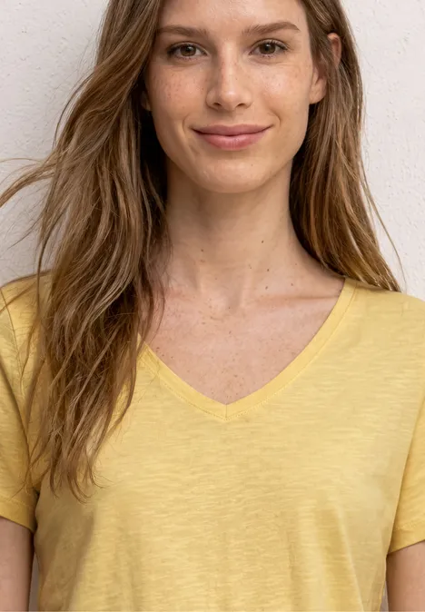 Vrouw in geel V-hals T-shirt met lange haren, voor een witte muur.