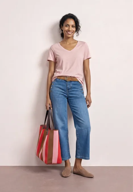 Frau in rosa T-Shirt, blauen Jeans und beigen Slippern trägt gestreifte Tasche vor hellem Hintergrund.