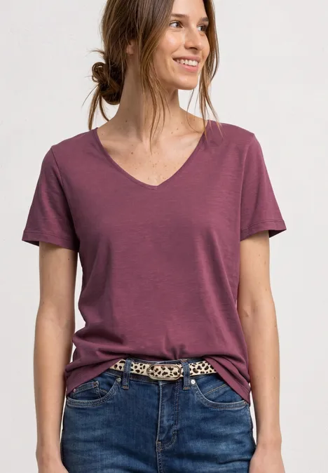 Vrouw in bordeaux T-shirt met V-hals en jeans met luipaardriem, haar in een knot.