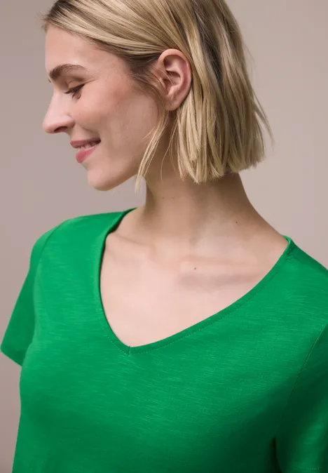 Persoon met halflang blond haar draagt een groen V-hals t-shirt tegen een neutrale achtergrond.
