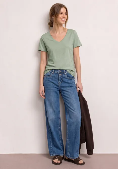 Person in grünem V-Ausschnittshirt und weiten blauen Jeans, trägt braune Sandalen und hält eine Jacke.