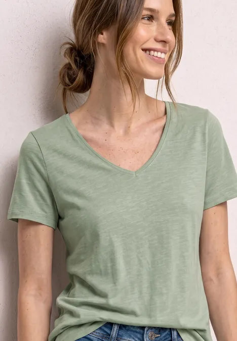 Frau in hellgrünem V-Ausschnitt-T-Shirt, lockerer Dutt, vor weißem Hintergrund.