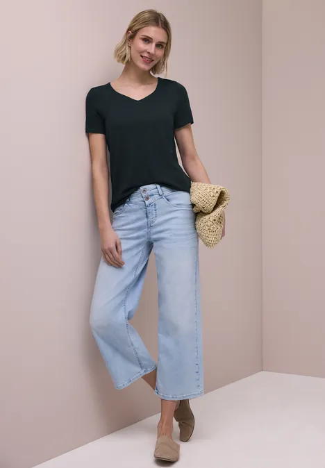 Vrouw in zwart T-shirt met lichtblauwe wijde jeans, strooien tas en beige sleehakken voor een neutrale achtergrond.