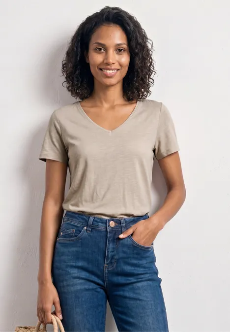 Basic T-Shirt mit V-Neck in Unifarbe light sepia sand Frau in beigem V-Neck-Shirt und blauen Jeans steht vor weißem Hintergrund und hält Tasche.
