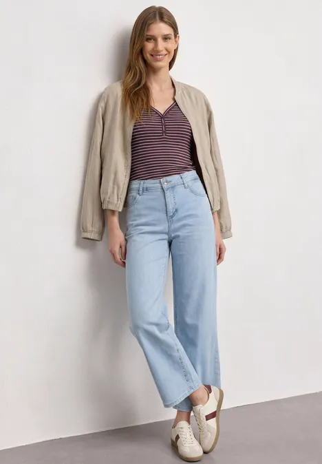 Top mit V-Neck und Streifen rustic rouge Eine Frau steht vor einer weißen Wand, trägt hellblaue Straight-Leg-Jeans, gestreiftes Shirt und beigen Blouson.