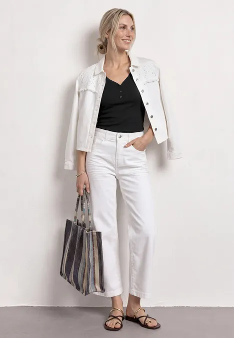 Frau mit weißer Jacke, schwarzem Top, weißen Jeans, Sandalen und gestreifter Tasche vor weißem Hintergrund.
