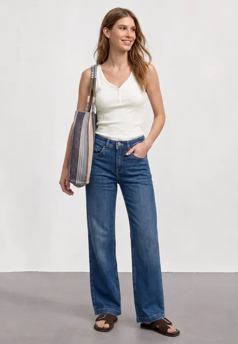 Eine Frau mit langen braunen Haaren trägt ein weißes Top, weite blaue Jeans, schwarze Sandalen, eine gestreifte Tasche.