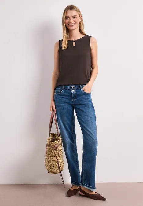 Frau in schwarzem ärmellosem Top, blauen Jeans und braunen Loafers mit geflochtener Strohtasche vor weißem Hintergrund.