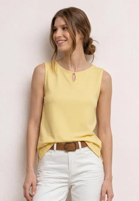 Vrouw in gele mouwloze top en witte broek met bruine riem, staand voor een lichte achtergrond.