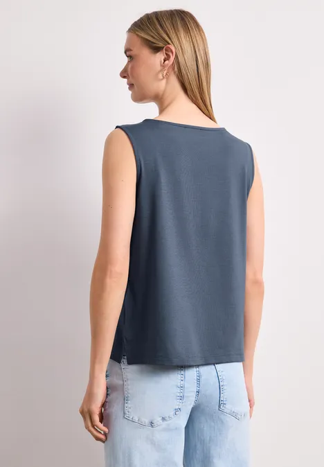 Rückansicht einer Frau mit schulterfreiem, dunkelgrauen Top und hellblauen Jeans vor neutralem Hintergrund.